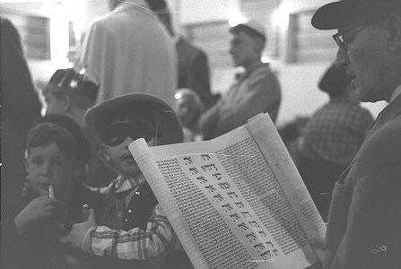 A Jewish man reads the Megillah (Book of Esther) during Purim, surrounded by children dressed in costumes for the festival. (Credit: GPO National Photo Collection)
